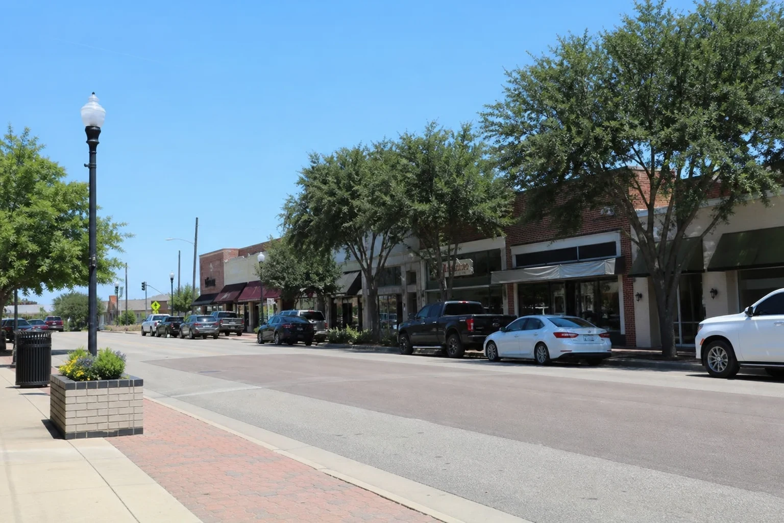 Sunny main street lined with brick storefronts, green awnings and shade trees, with pickups and sedans parked along the curb beside a brick-paved sidewalk