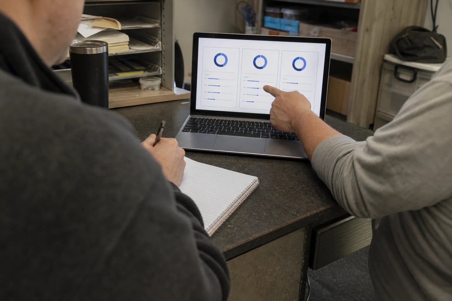 Over-the-shoulder view of a man taking notes on a spiral notepad while another person points at three chart options on a laptop at a cluttered office desk