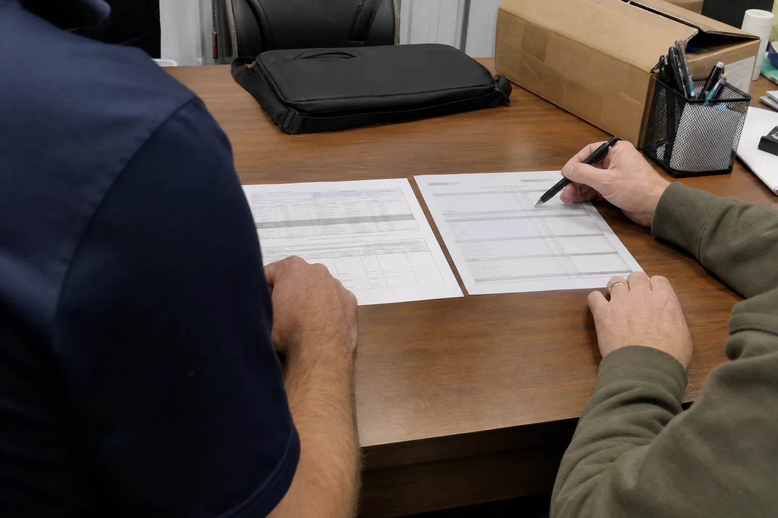 Two people at a wooden office desk comparing two printed forms laid side by side, one pointing with a pen, with a laptop bag, cardboard box and pen holder nearby