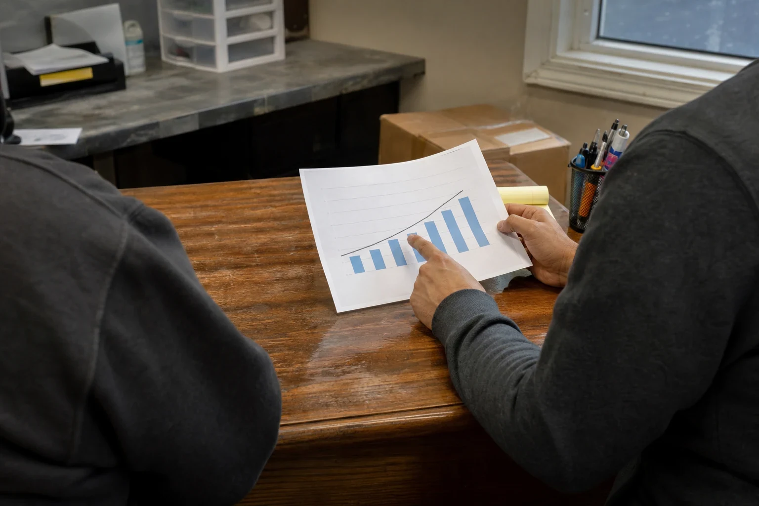 Two people seated at a wooden desk in a small back office, one pointing at a printed bar chart while holding the page, with boxes and a pen cup nearby